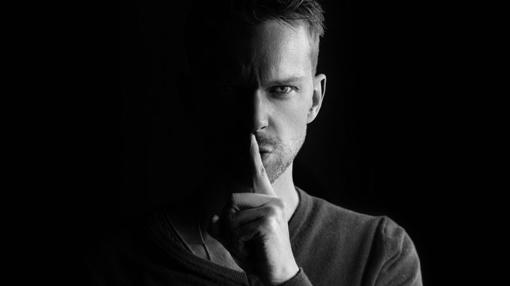 Black and white photo of an adult man holding his finger up to his mouth to indicate a secret